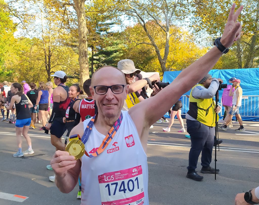 Uśmiechnięty autor artykułu, Witold Tarnowski, stoi w słonecznym Central Parku tuż po ukończeniu maratonu. Ubrany jest w białą koszulkę startową z godłem Polski, logo Orlen i numerem startowym 17401. W prawej dłoni dumnie trzyma duży, złoty medal, a lewą rękę ma uniesioną w górę, pokazując palcami znak zwycięstwa (V). W tle widać jesienne drzewa i innych uczestników imprezy.