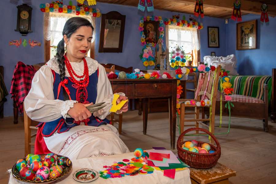 Kobieta w tradycyjnym stroju ludowym wycinająca ozdoby z papieru w wiejskiej izbie, obok koszyka z kolorowymi pisankami