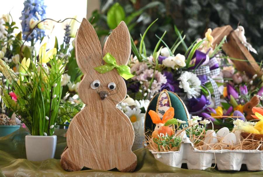 Drewniana figurka wielkanocnego zająca z zieloną kokardką na tle kolorowych dekoracji, kwiatów i pisanek.
