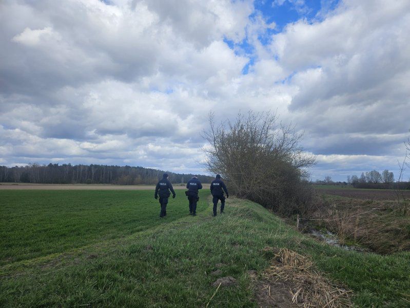 Trzej policjanci w ciemnych mundurach idący skrajem zielonego pola wzdłuż zarośli pod zachmurzonym niebem