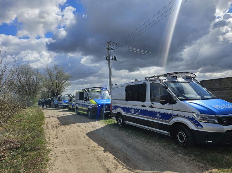 Szereg radiowozów polskiej Policji typu furgon zaparkowanych na polnej drodze pod zachmurzonym niebem
