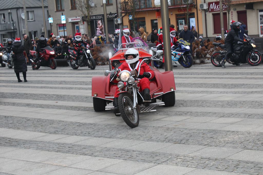 Motocyklista przebrany za Świętego Mikołaja prowadzi czerwony, trzykołowy motocykl (trike) po wybrukowanym placu miejskim. Wokół niego jadą inni motocykliści, również w strojach Mikołajów, a w tle widać zgromadzonych widzów i budynki w centrum miasta.