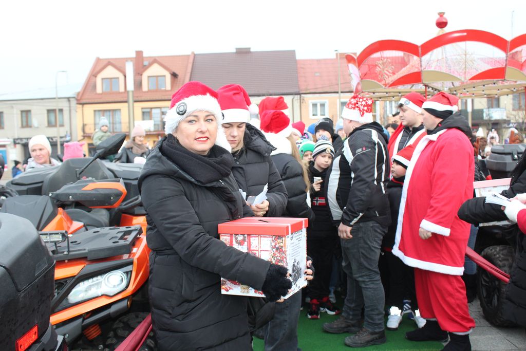Uśmiechnięta kobieta w czapce Świętego Mikołaja trzyma czerwone pudełko, prawdopodobnie na datki. Stoi obok pomarańczowego quada, a za nią znajduje się grupa ludzi, w tym dzieci i osoby przebrane za Mikołajów. W tle widać świąteczne dekoracje i karuzelę.