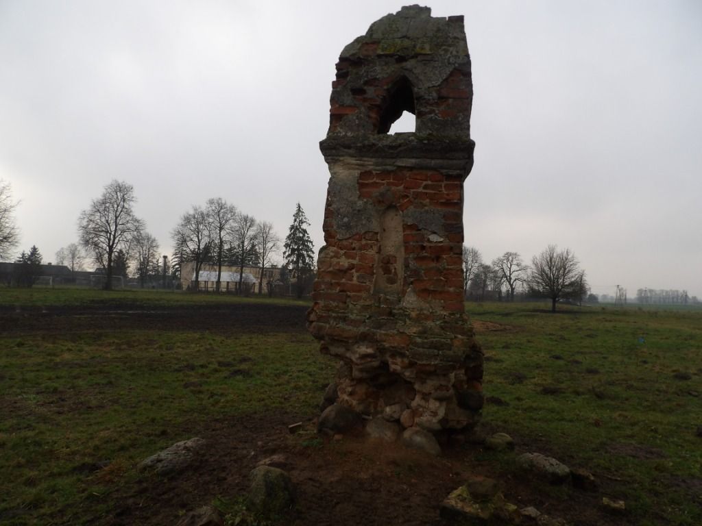 Zrujnowana, murowana kapliczka w kształcie słupa (obelisku) z czerwonej cegły i kamieni, stojąca na zielonej łące w Kowalewie na tle bezlistnych drzew w pochmurny dzień.