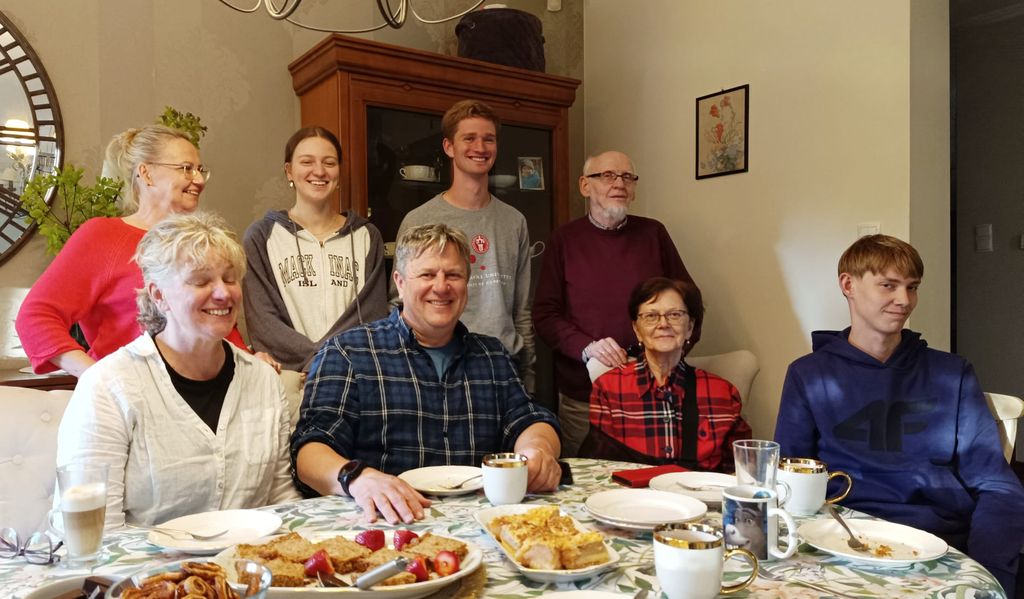 Grupowe zdjęcie ośmiu uśmiechniętych osób przy stole w jasnym salonie. Na stole widać filiżanki z kawą, talerze z ciastem i truskawkami. Wśród obecnych są osoby starsze oraz młodzież, wszyscy ubrani w stroje codzienne, patrzą w obiektyw.
