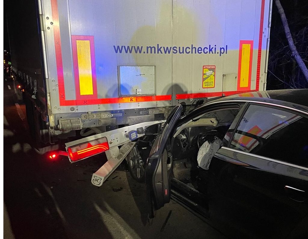 Rozbite osobowe Audi A6 stojące za naczepą samochodu ciężarowego na drodze wojewódzkiej w Płocochowie. W tle widoczne światła policyjnego radiowozu oraz sygnalizacja świetlna.