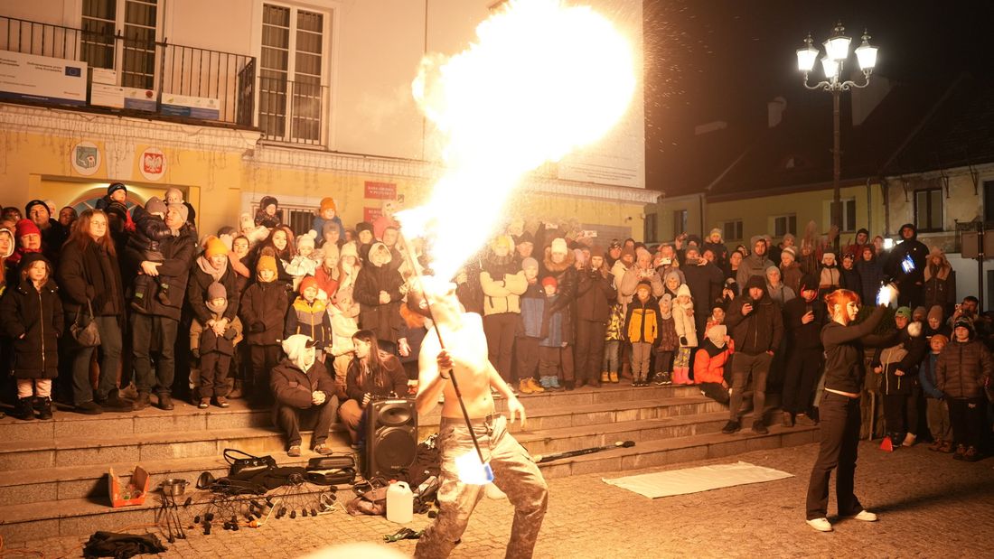 Nocny pokaz fire show na rynku miejskim. Artysta na pierwszym planie zionie potężnym słupem ognia, oświetlając zgromadzony na schodach tłum widzów w zimowych ubraniach.